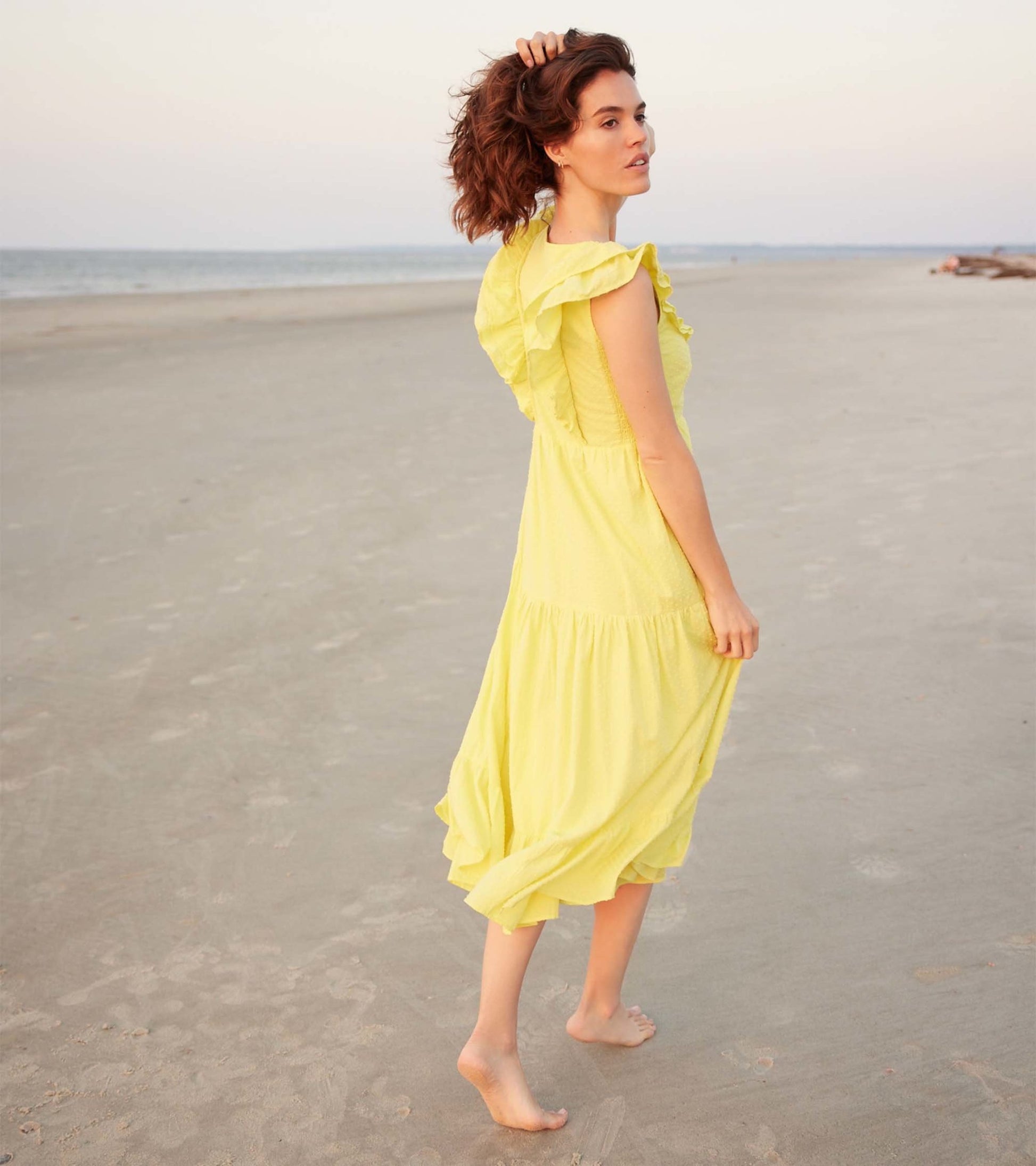 Yellow Swiss dot midi dress with ruffled sleeves, worn barefoot on a beach at sunset.