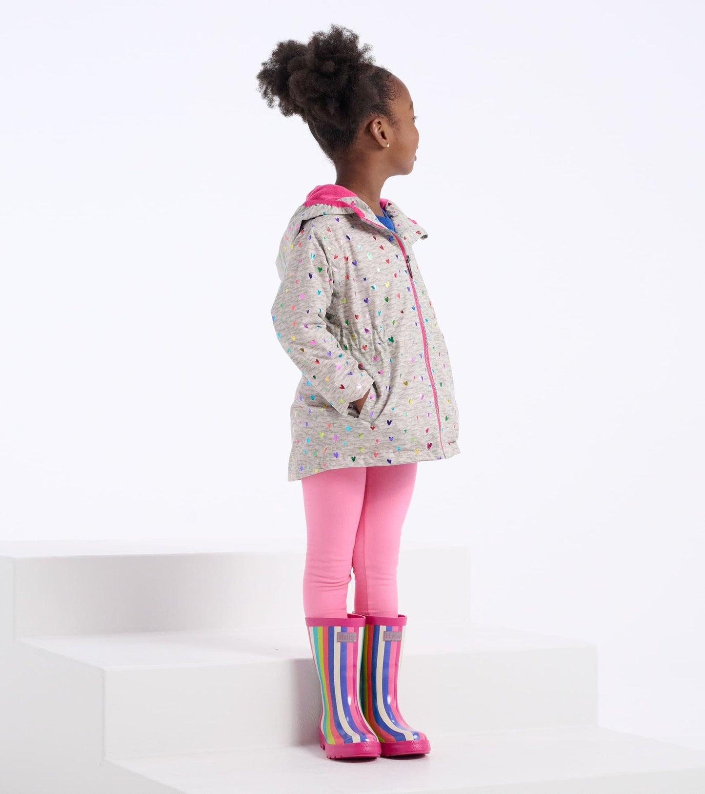 Lightweight zip-up rain jacket with colorful confetti hearts, paired with pink leggings and striped rain boots.