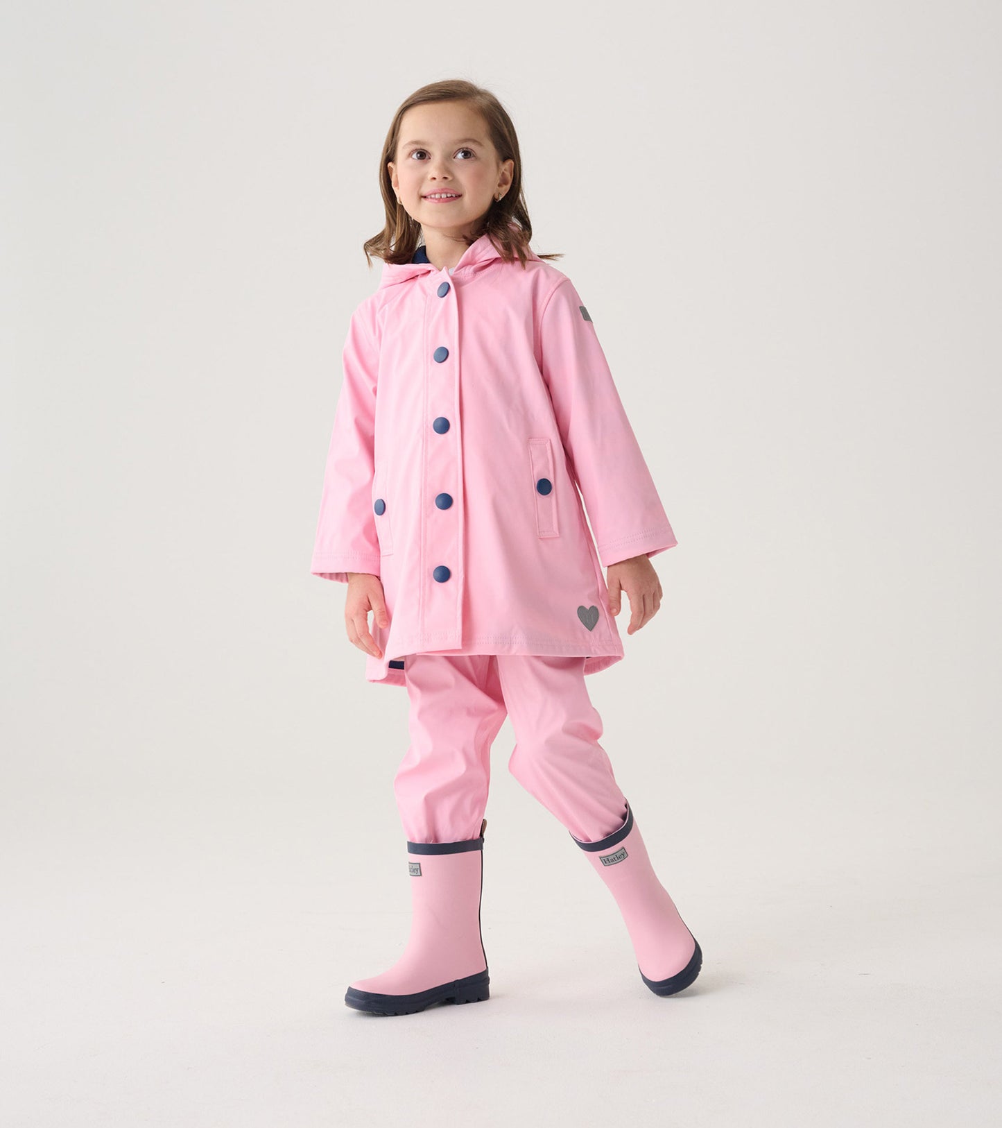 Pink button-up rain jacket with navy buttons, hood, and heart detail, worn by a girl with matching pink rain boots.