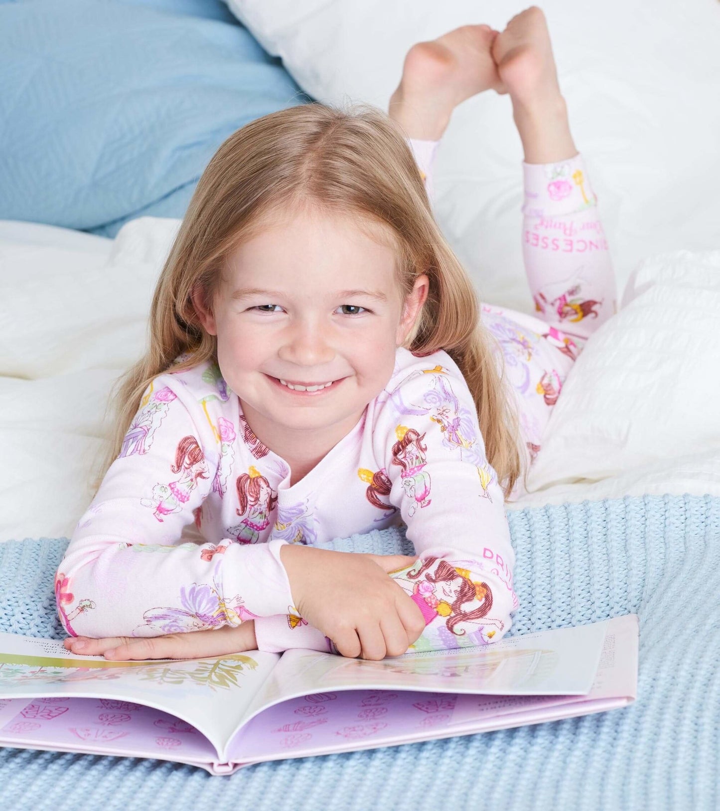Light pink pajama set featuring princess illustrations, worn by a smiling child reading on a bed with blue and white bedding.