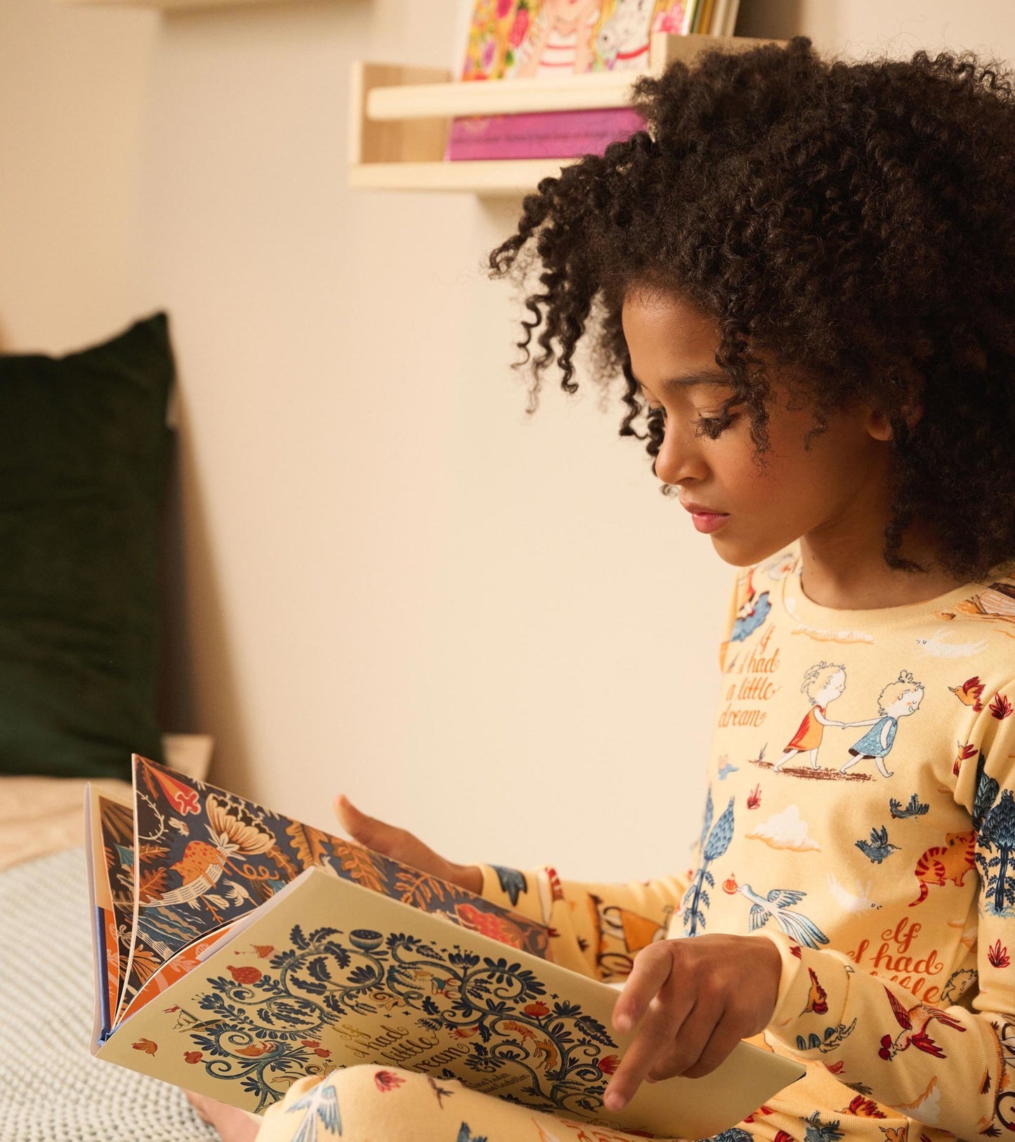 Child in yellow pajama set with whimsical patterns, reading a colorful storybook while seated on a bed.