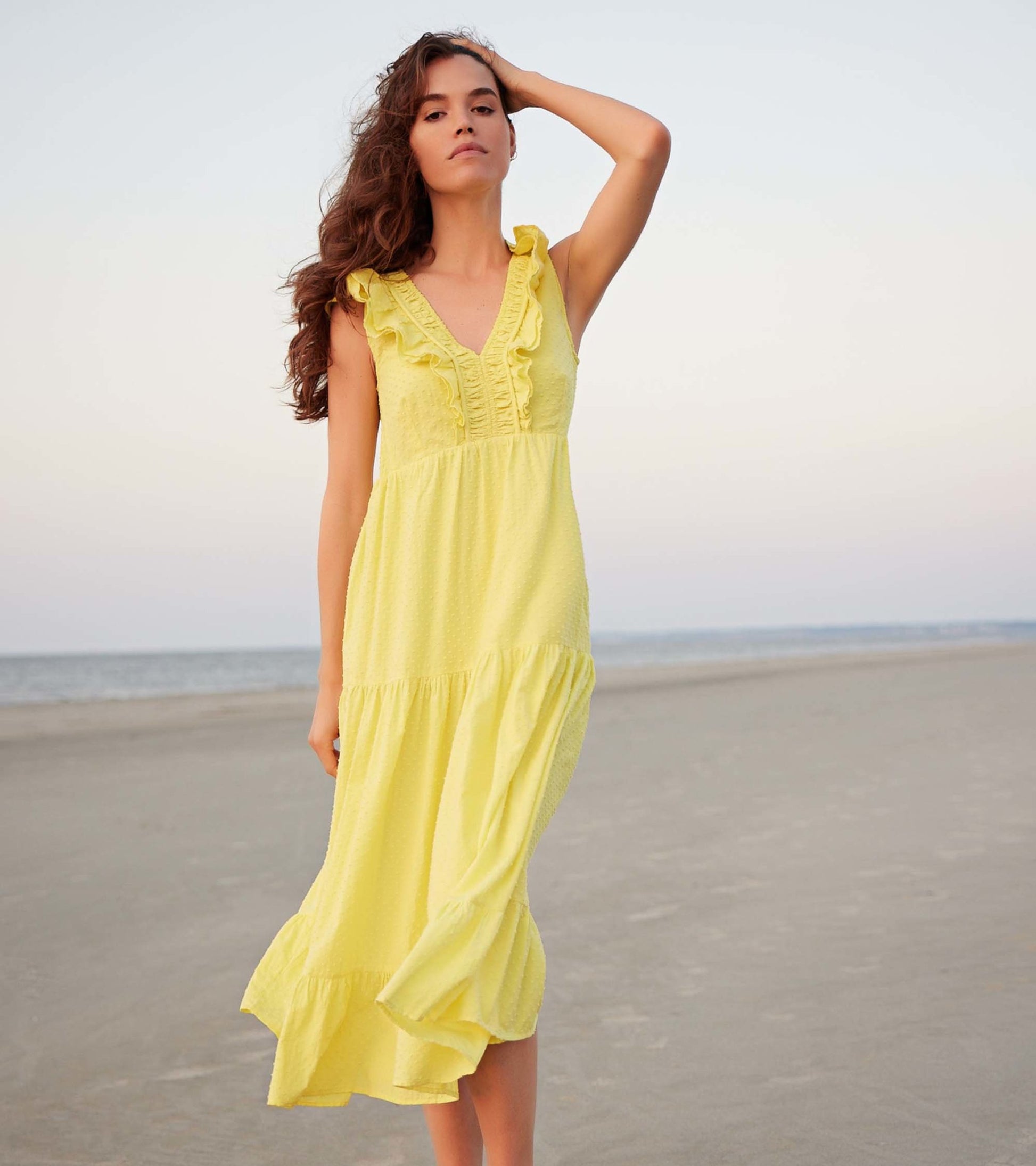 Yellow Swiss dot midi dress with ruffled straps and a tiered skirt, worn by a model on a beach.