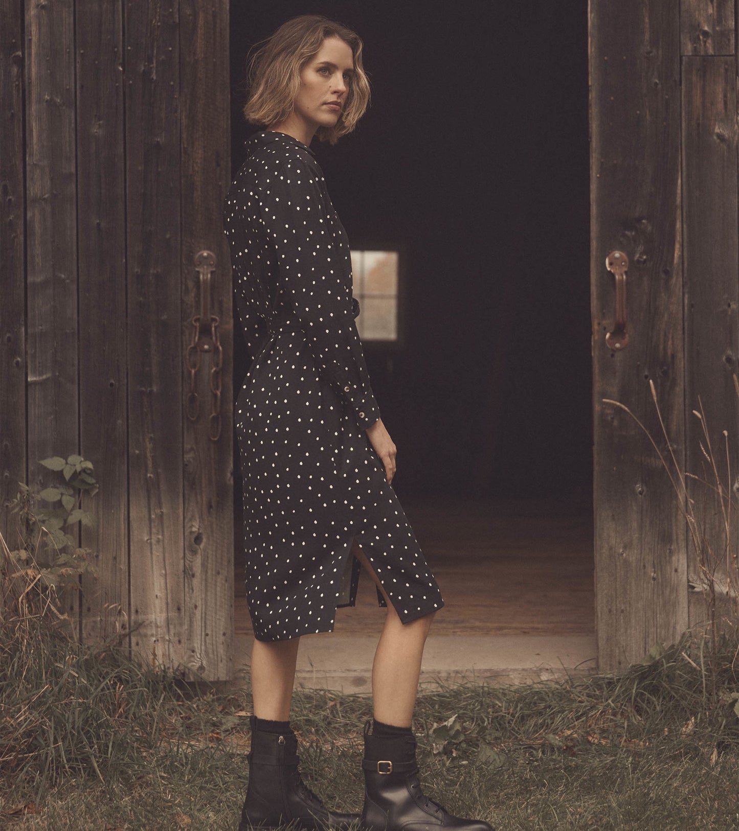 Black midi shirt dress with white polka dots, featuring a front slit and long sleeves, styled with black ankle boots.