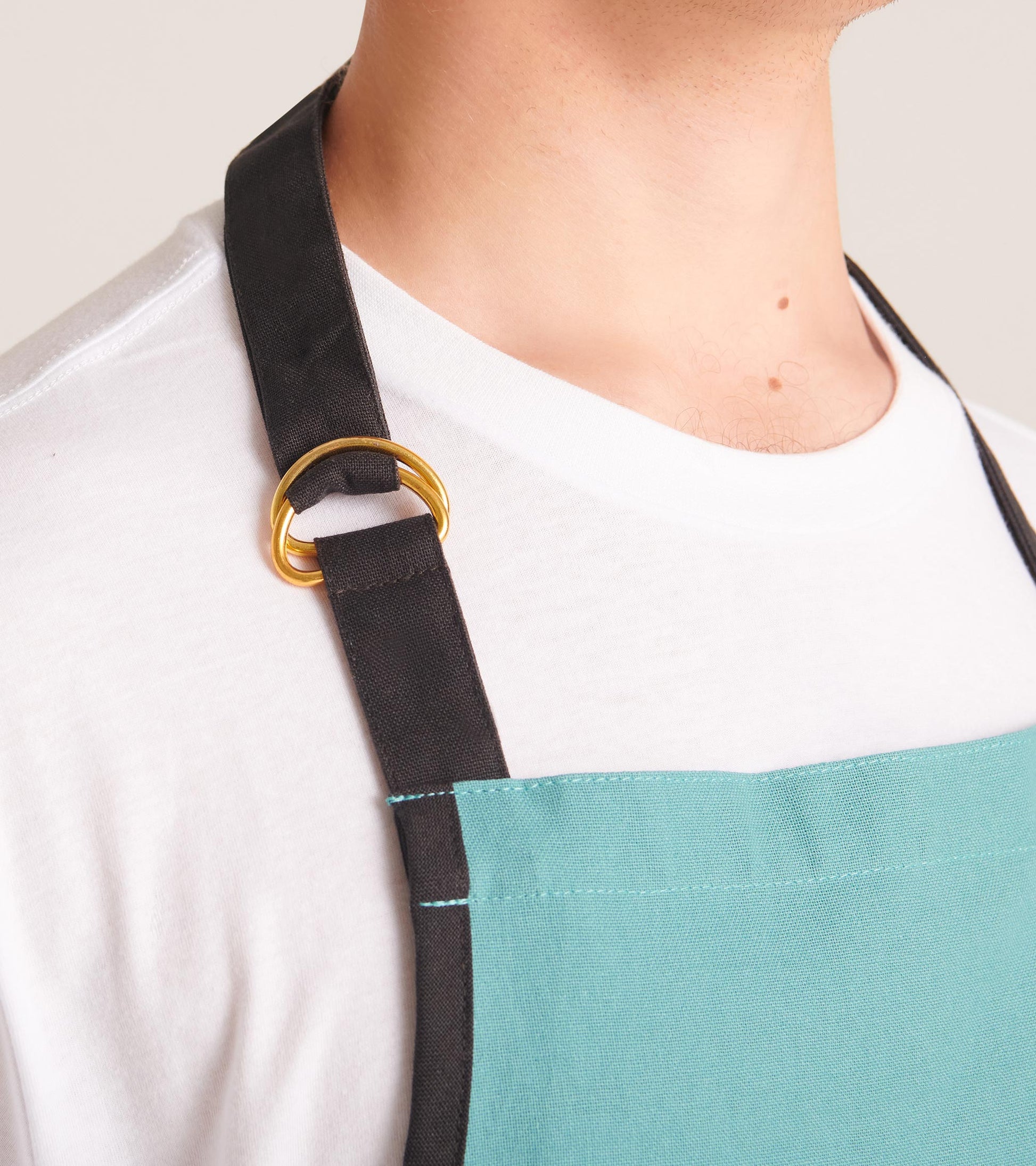 Close-up of a white apron with black neck strap, gold ring adjuster, and turquoise front panel