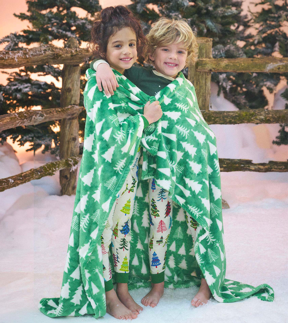 Two children wrapped in a green fleece blanket with white Christmas tree pattern