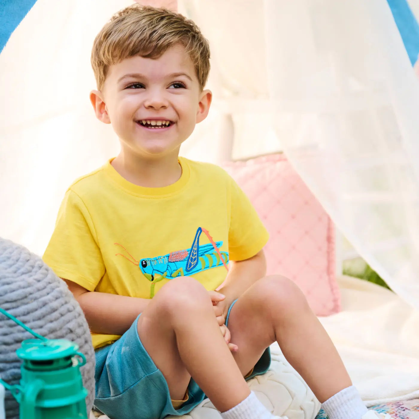 Promotes baby and kids clothing. Showing smiling young boy in a yellow graphic t-shirt and blue shorts seated indoors.