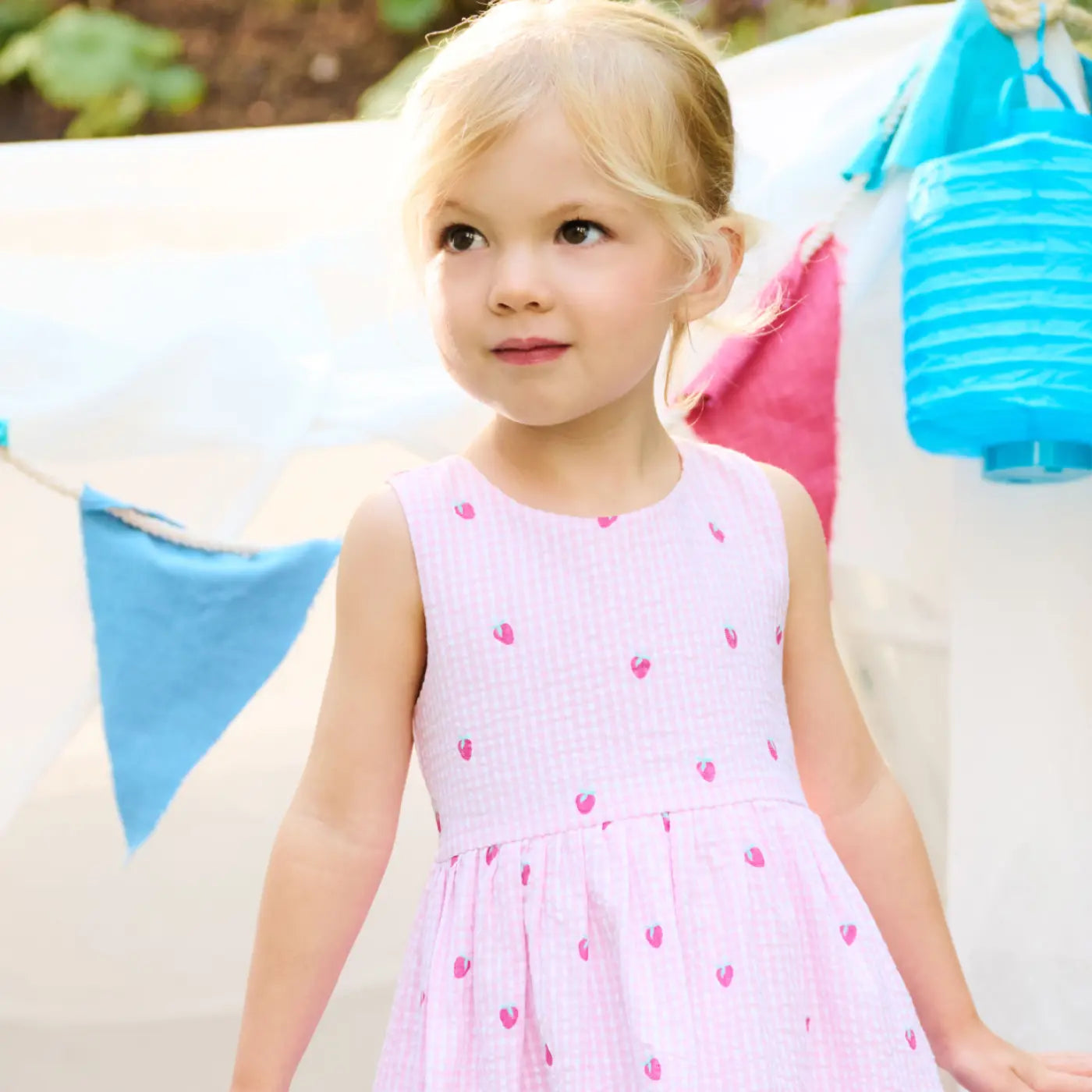 Promotes baby and toddler clothing. Showing young girl in a light pink sleeveless dress outdoors with colorful lanterns.