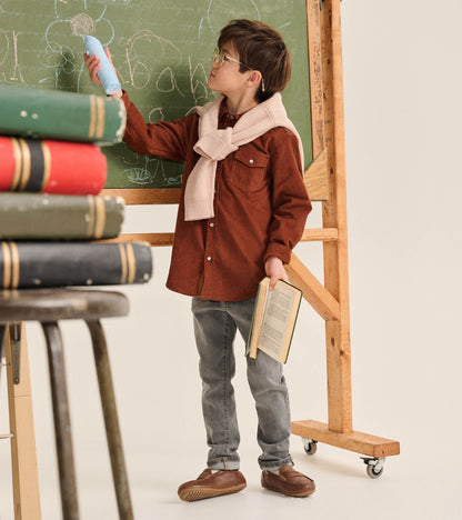 Boy wearing charcoal grey jeans with a brown shirt, standing by a chalkboard and holding a book