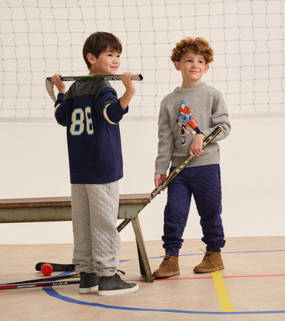 Boy wearing patriot blue quilted joggers in a gym, paired with a gray sweater and brown lace-up boots