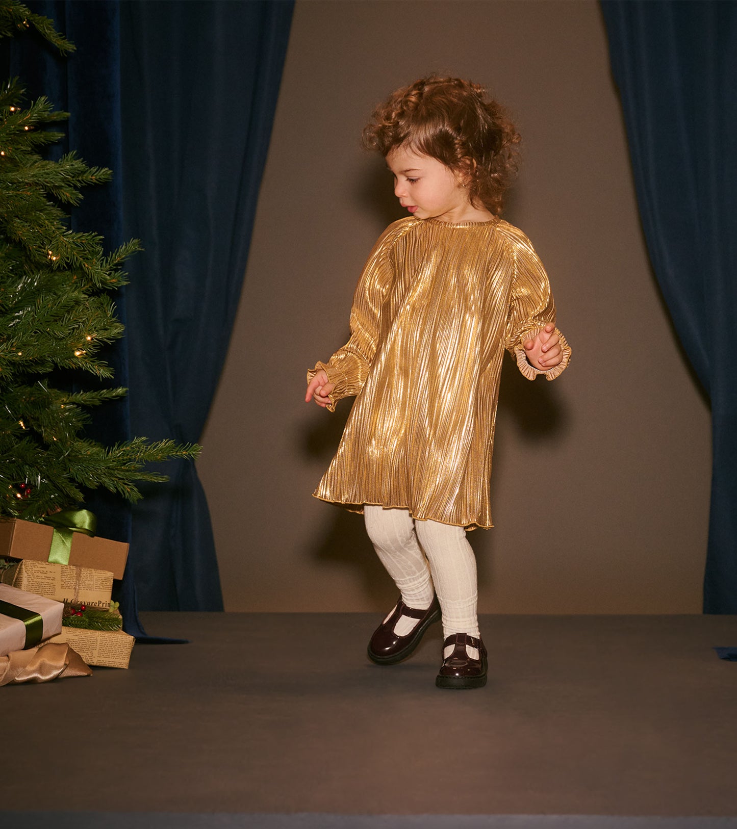 Toddler wearing a gold metallic pleated dress with long sleeves in a holiday scene, front view