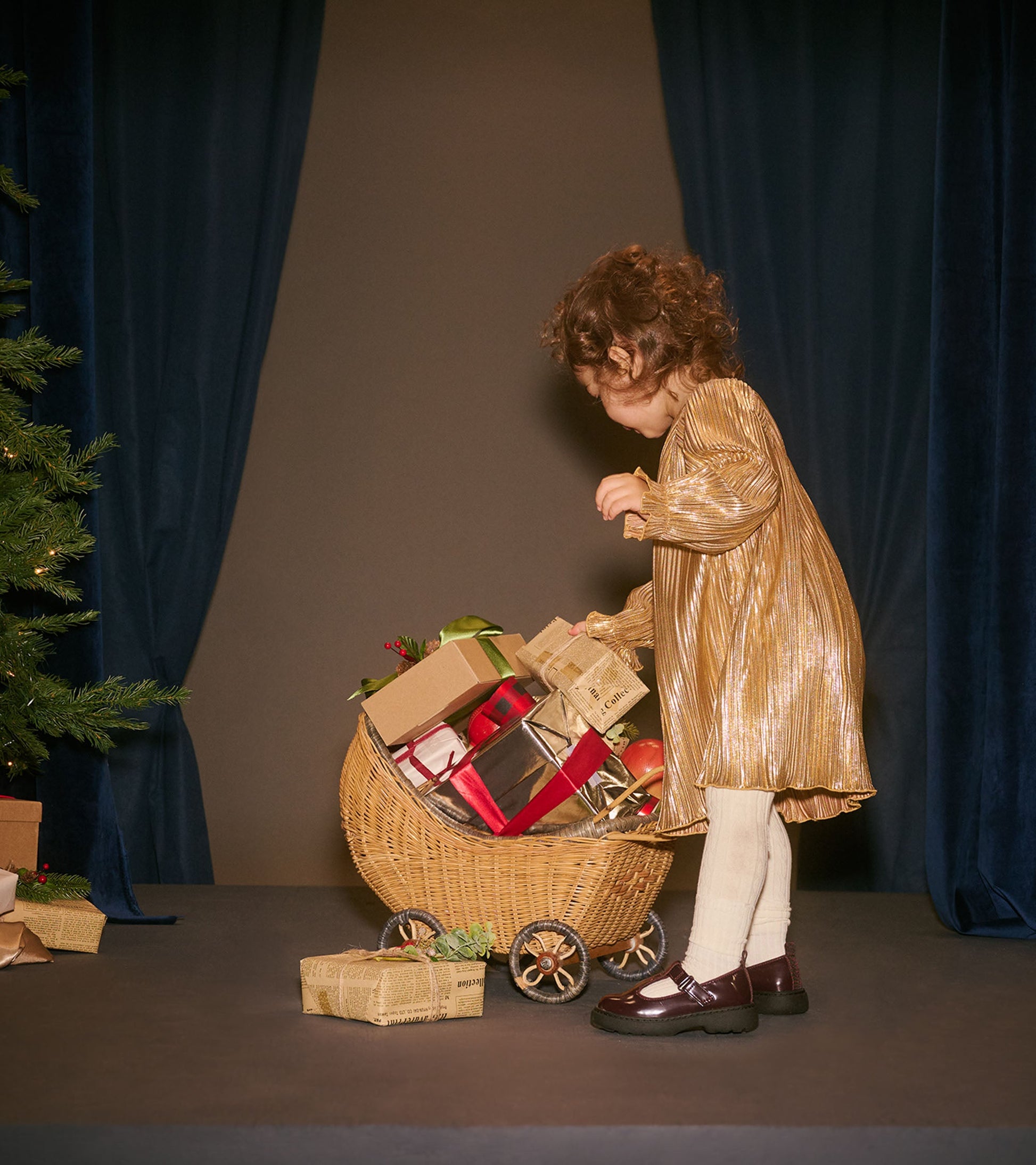 Gold metallic special dress with long sleeves, worn by a toddler, while playing with a cart of wrapped gifts.