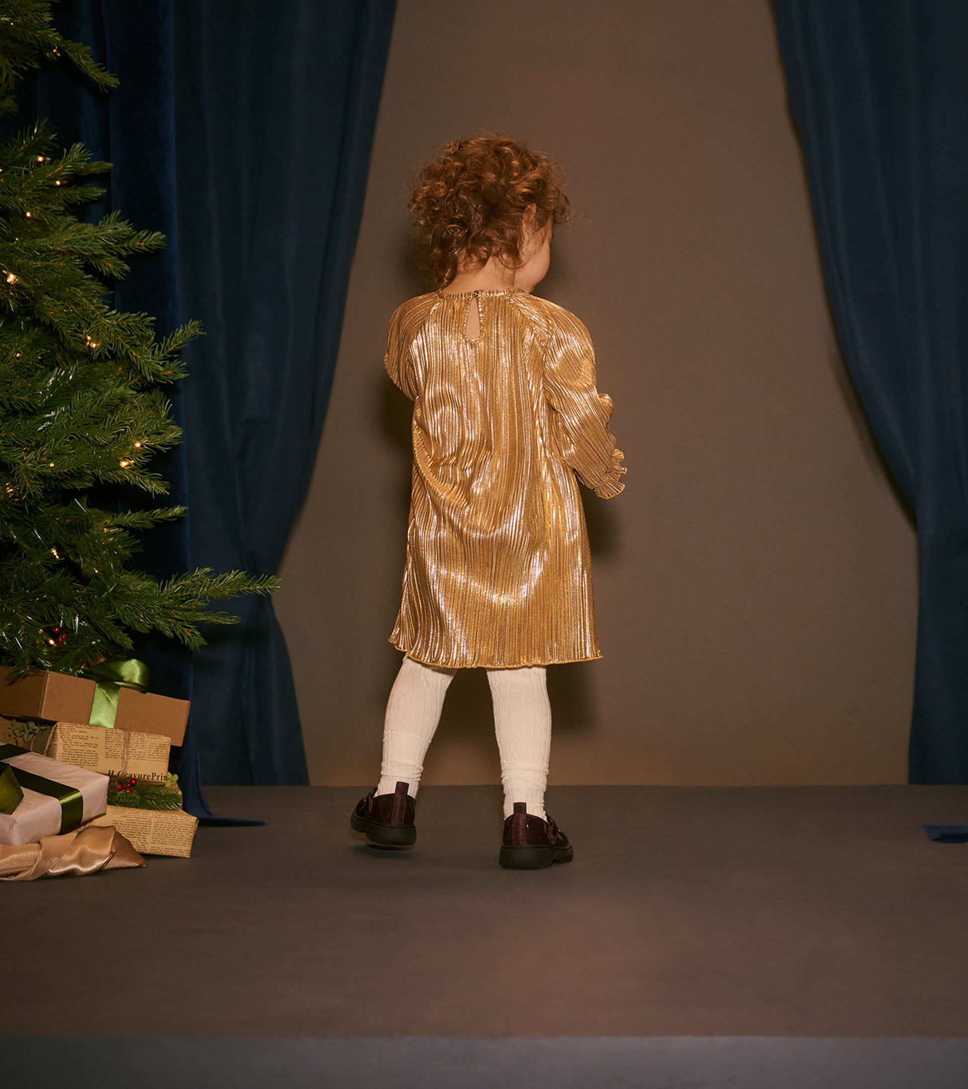 Toddler wearing a gold metallic pleated dress in a holiday scene, back view