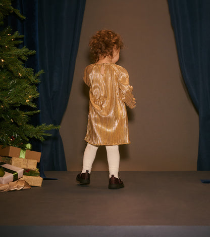 Toddler wearing a gold metallic pleated dress in a holiday scene, back view