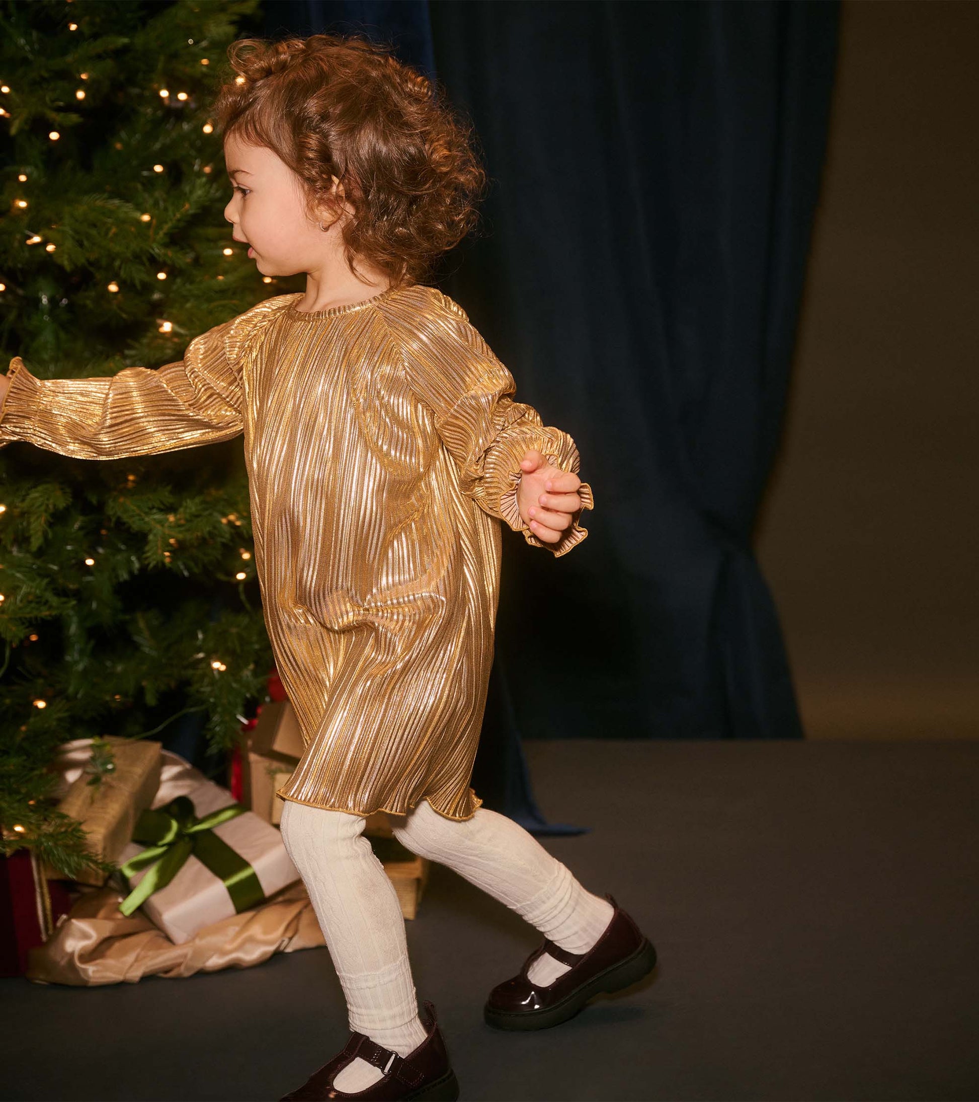 Gold metallic special dress with long sleeves and ruffled cuffs, worn by a toddler, paired with white tights and black shoes.