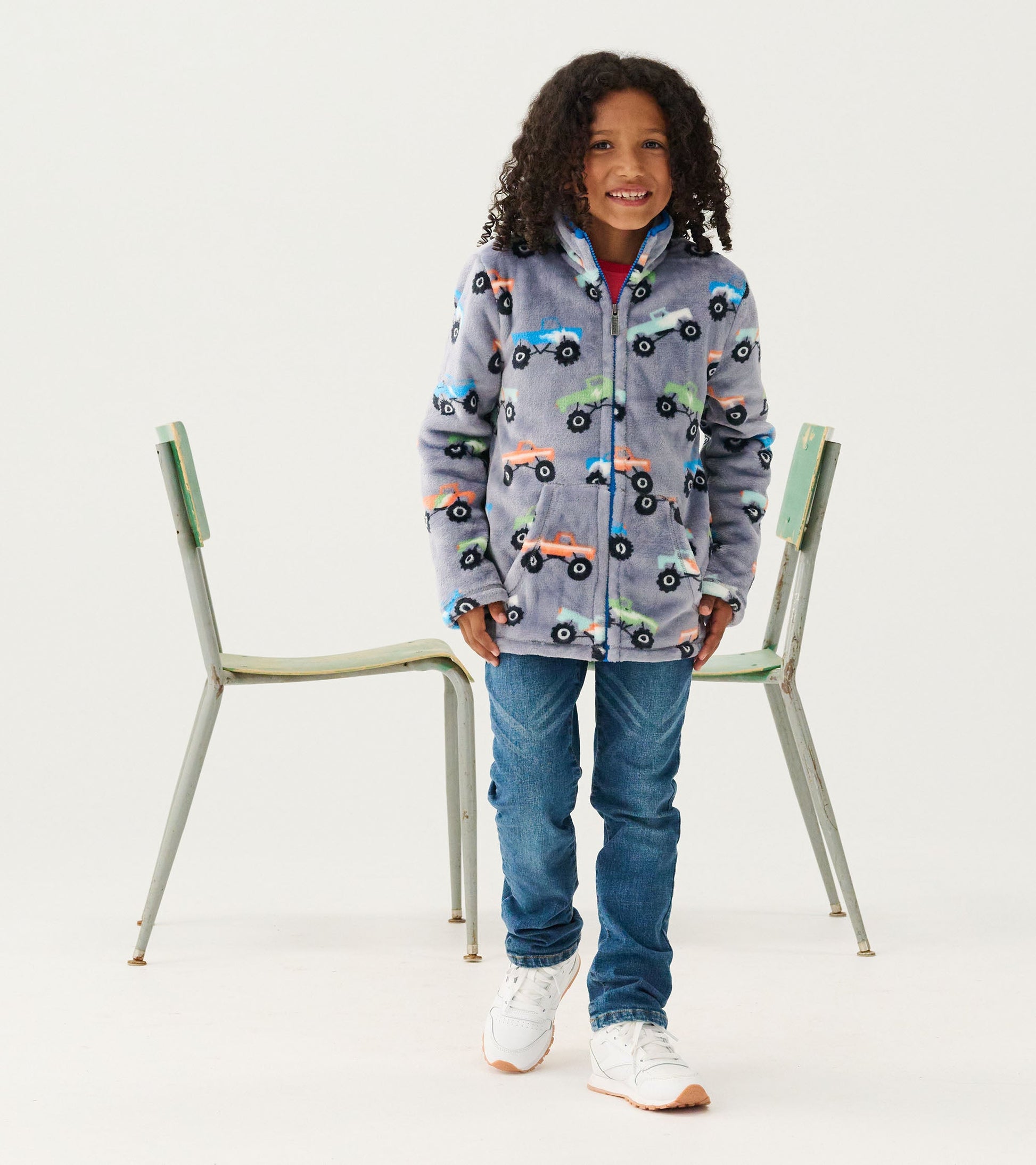 Fuzzy fleece zip-up jacket in light gray with colorful monster truck patterns, worn by a child standing in front of a chair.