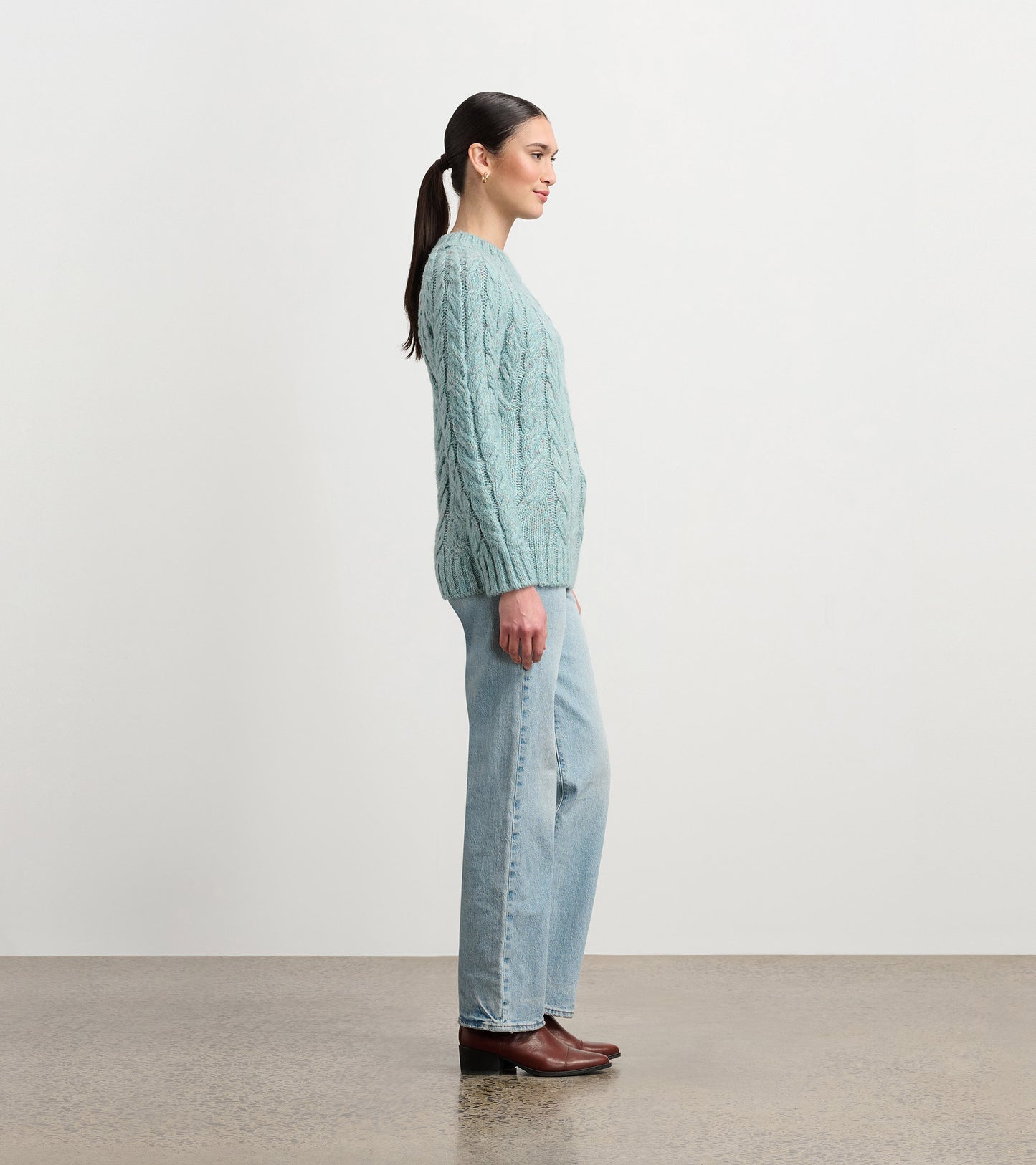 Light turquoise cable knit sweater with a relaxed fit, worn with light blue jeans and brown boots, shown from the side.