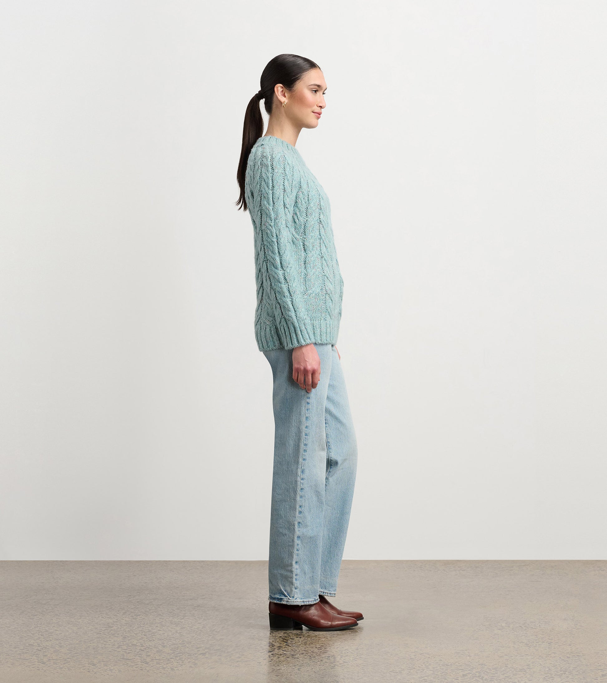 Light turquoise cable knit sweater with a relaxed fit, worn with light blue jeans and brown boots, shown from the side.