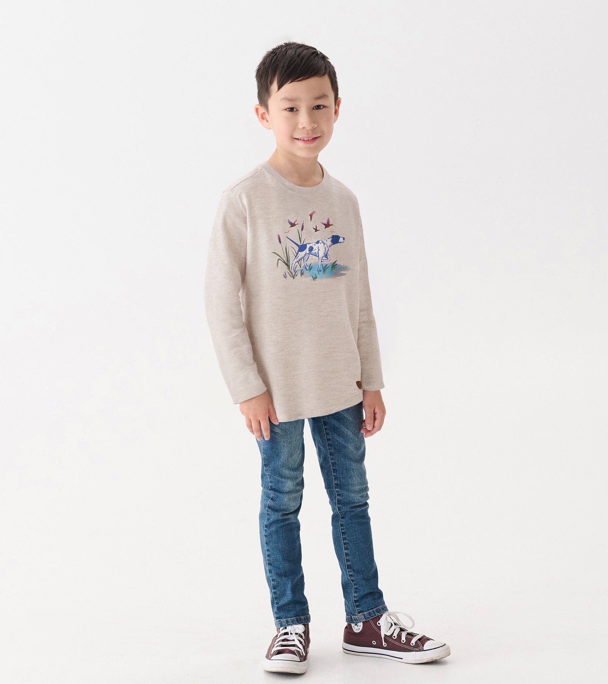Light beige long sleeve tee featuring a graphic of a hunting dog among flowers, paired with blue jeans and sneakers.