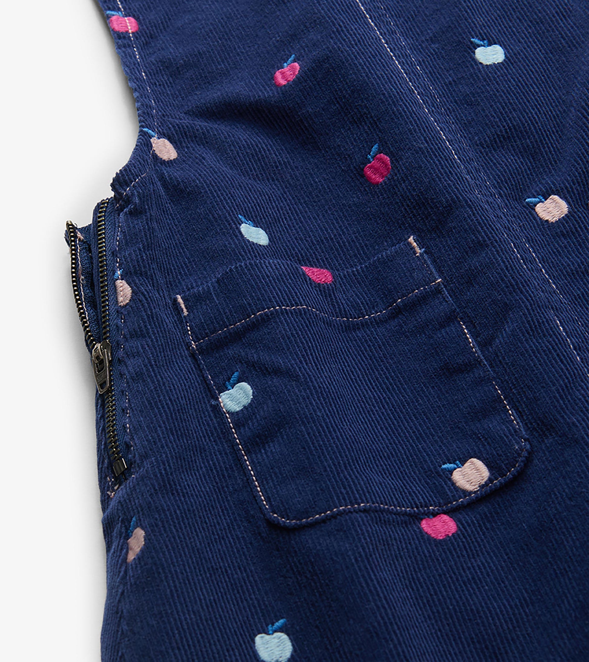 Close-up of navy pinafore dress showing embroidered apples, front pocket, and side zipper