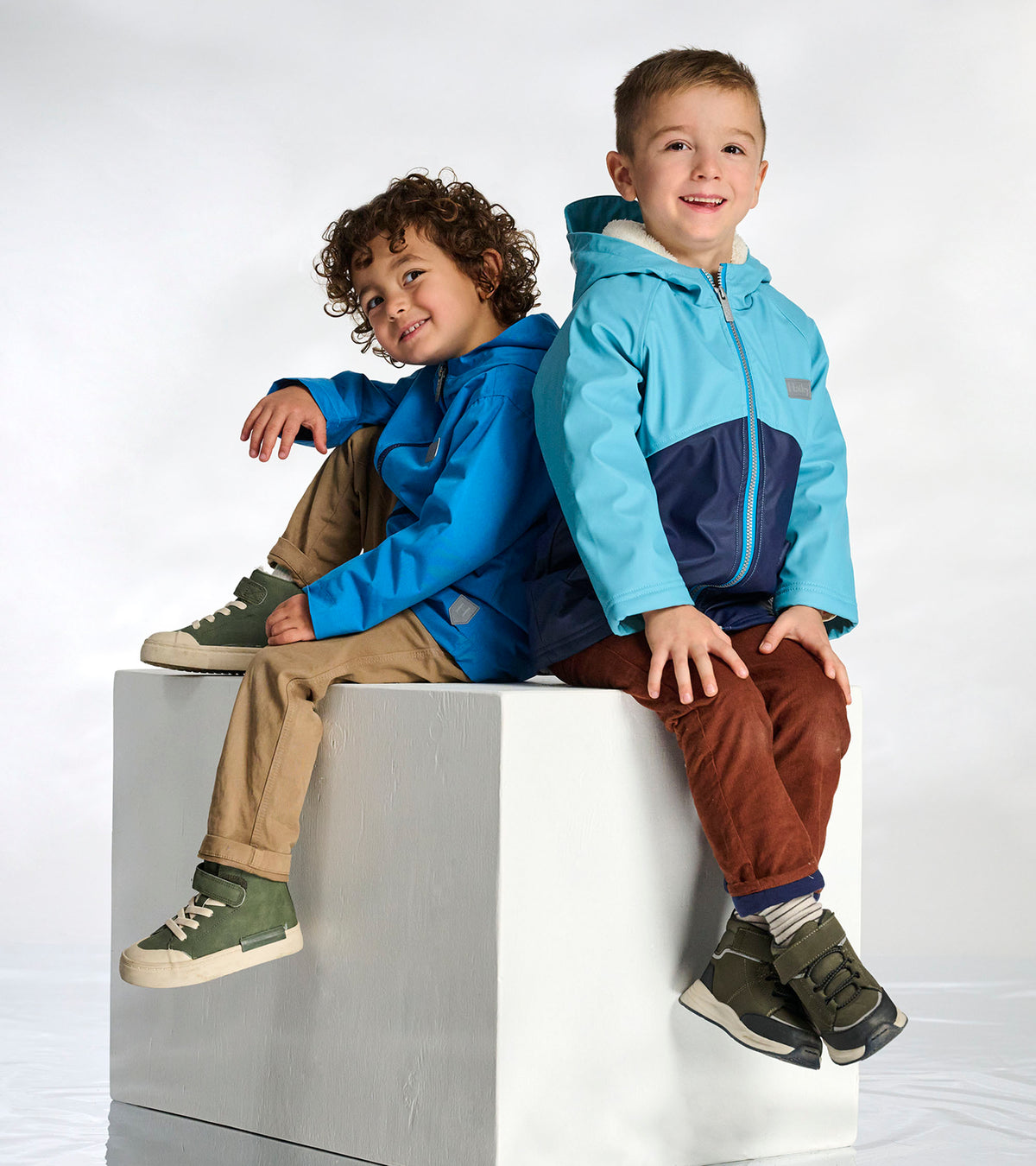 Boy wearing blue colourblock sherpa-lined rain jacket seated indoors beside another child in a blue jacket