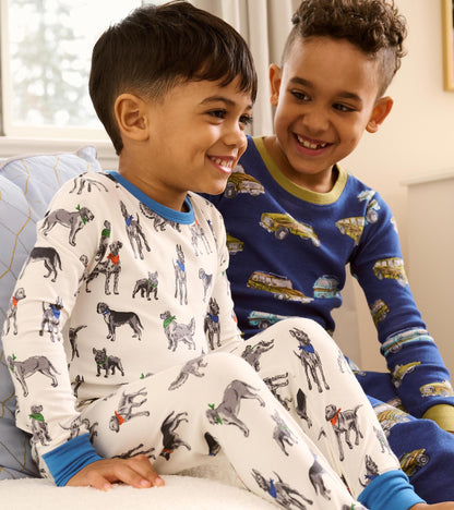 Child wearing white boys pajama set with grey dog print and bright blue trim, seated with another child
