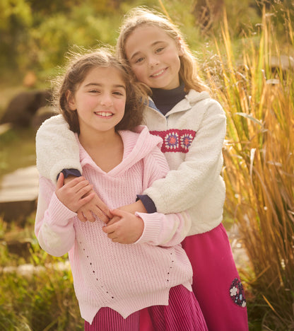 Girl wearing a cream fuzzy pullover with crochet chest band, hugging another child outdoors