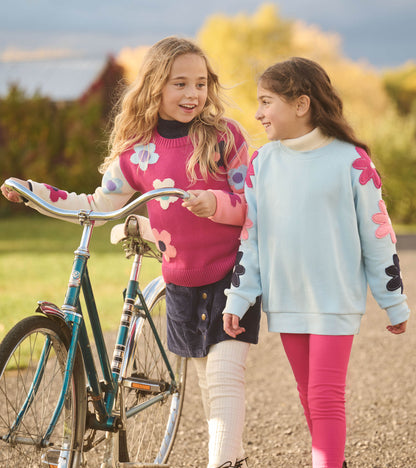 Girl wearing a cool blue pullover with pink and navy flower appliqués on the sleeves outdoors