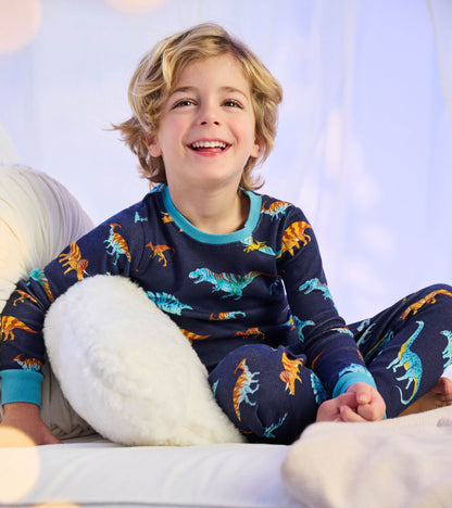 Child wearing navy dinosaur print pajamas with teal neckline and cuffs, seated on a bed