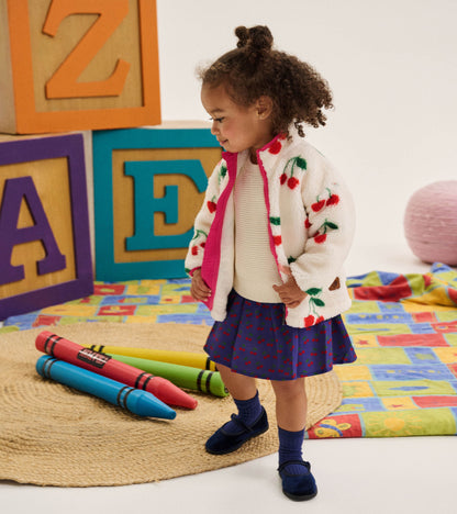 Side view of toddler wearing the cream knit and purple cherry-print sweater dress under a cherry jacket