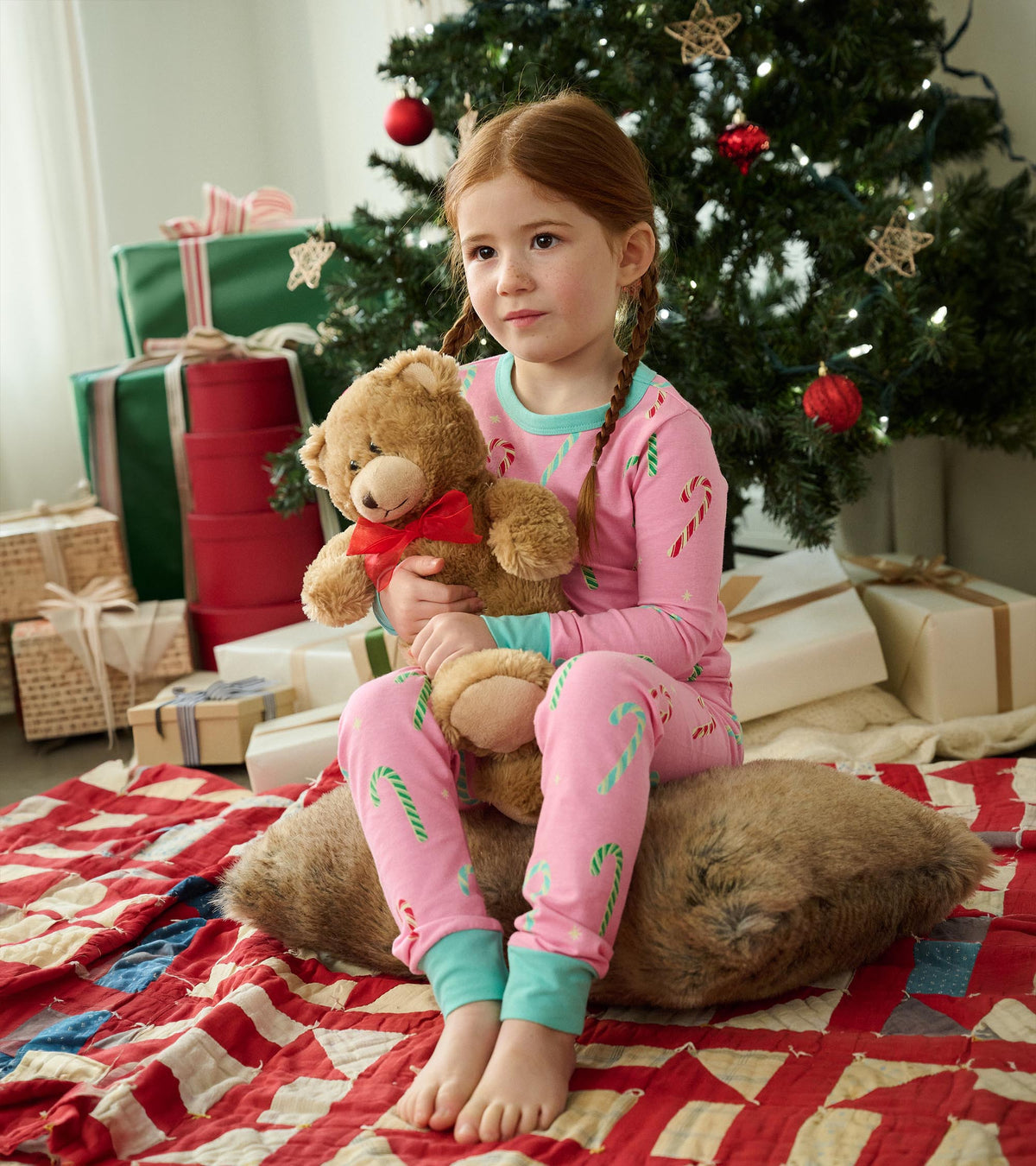 Girl wearing pink candy cane print pajamas with mint cuffs, seated by a Christmas tree holding a teddy bear