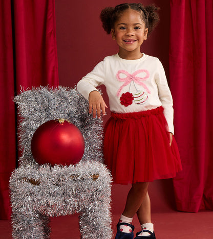 Toddler wearing a red puffy tutu skirt with a white holiday top beside silver tinsel decor