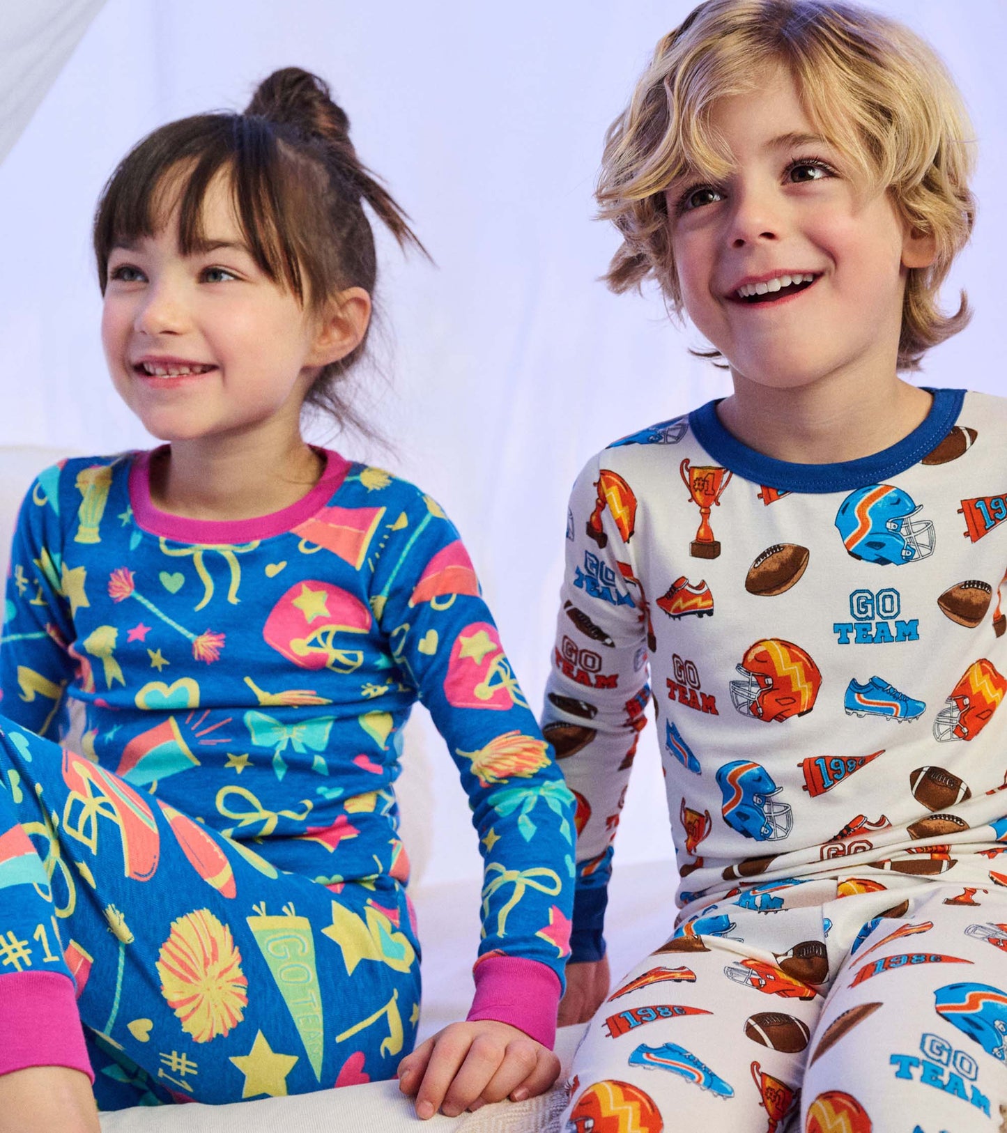 Girl wearing bright blue cheer print pajamas with pink trim, seated beside child in white sports print pajamas