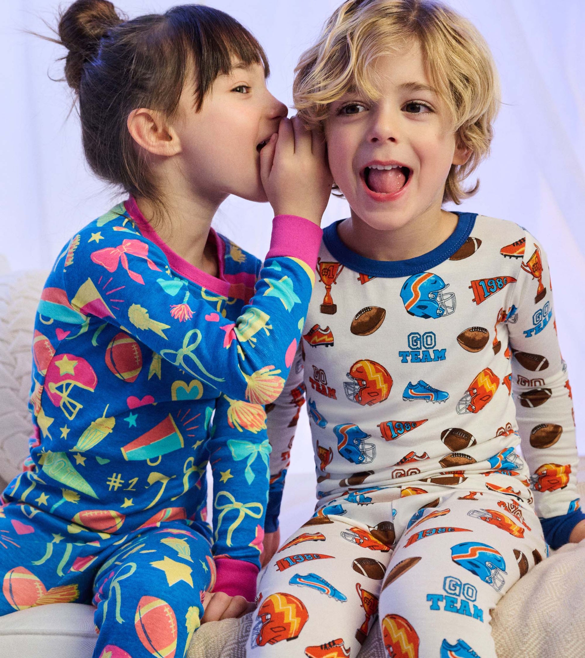 Girl in blue cheer print pajamas whispers to child in white sports print pajamas while sitting on a bed