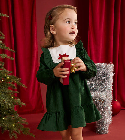 Toddler wearing an eden green ruffle dress with white bib collar and red bow, holding a red gift box
