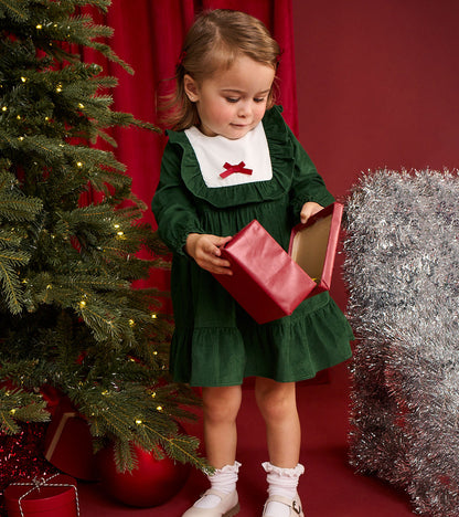 Toddler modeling green ruffle dress with white bib collar and red bow beside a Christmas tree