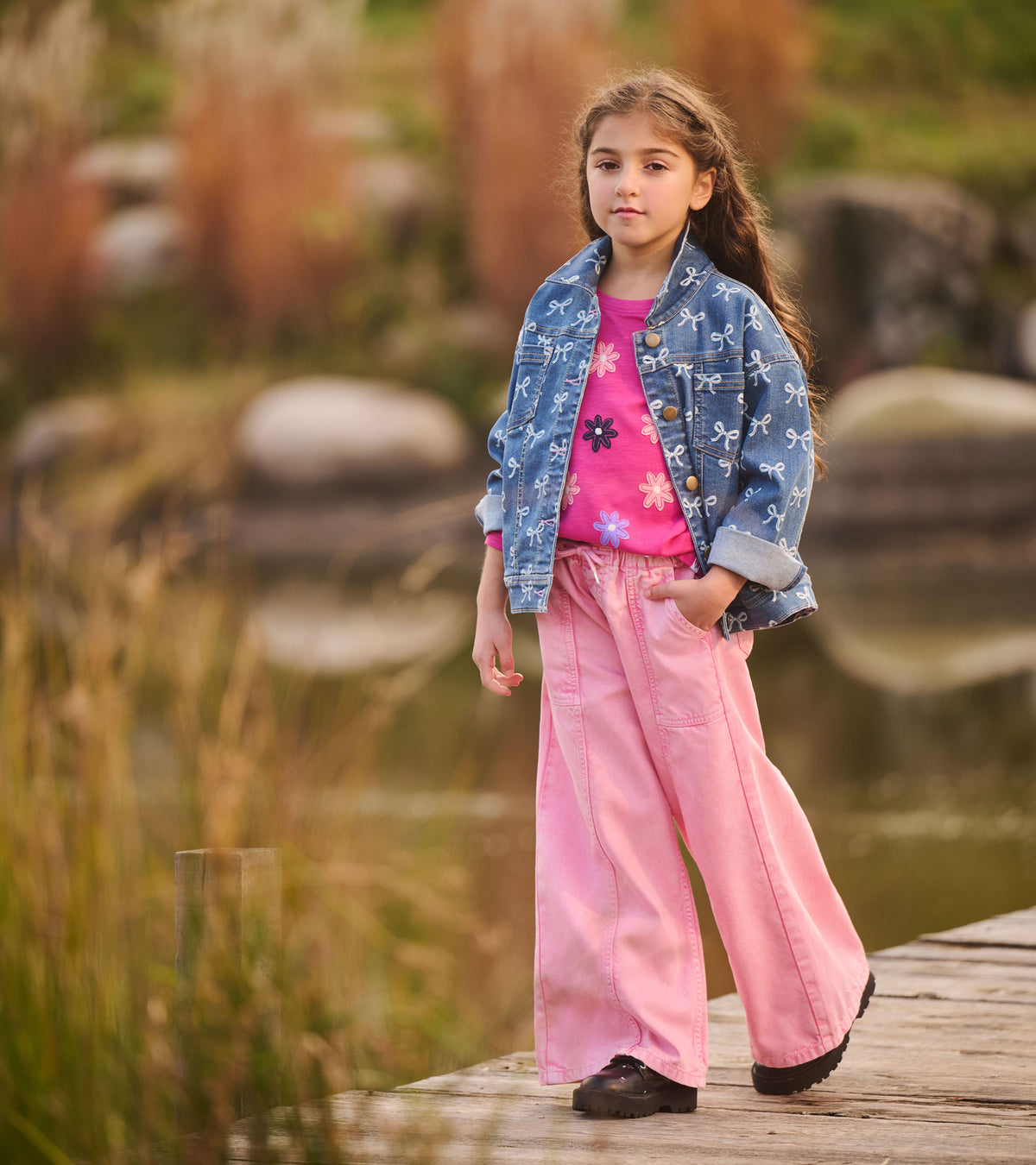 Girl wearing flamingo pink wide leg jeans with a blue bow-print jacket on a wooden path