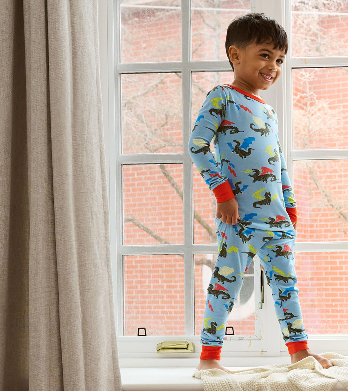 Child wearing blue dragon print pajamas with red cuffs, standing by a window