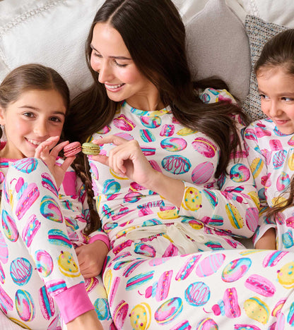 Girl wearing white pajamas with pink, yellow and blue macaron print, seated with family on a bed
