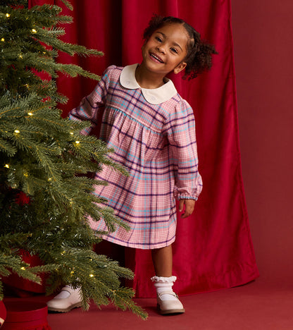Toddler girl wearing a mauve plaid long sleeve dress with cream Peter Pan collar beside a lit Christmas tree