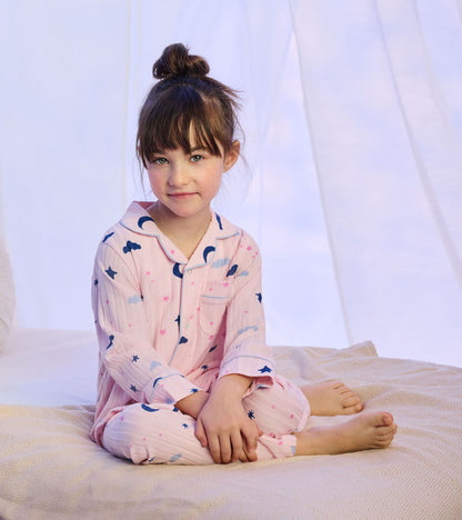 Girl wearing pink pajama set with moon, star, and cloud print, sitting cross-legged on a bed