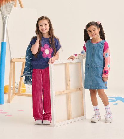 Girl wearing fuzzy blue flower sweater vest next to another child in a denim pinafore dress
