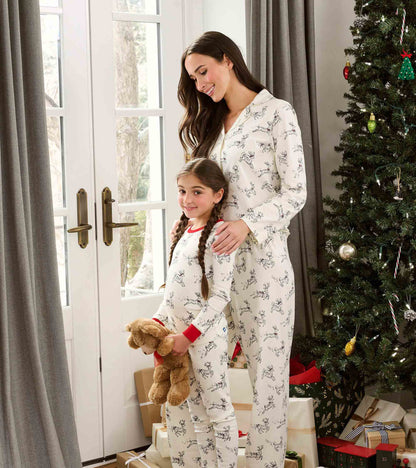 Girl in cream reindeer print pajamas with red trim standing by a Christmas tree, holding a teddy bear