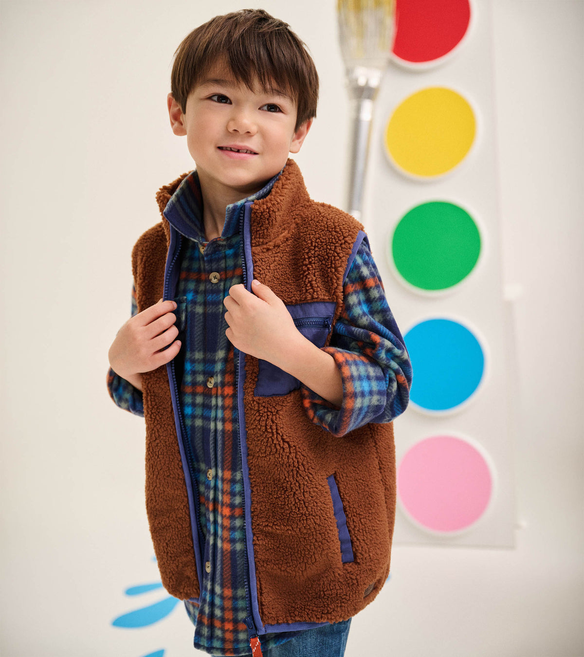 Boy wearing a brown sherpa vest with blue trim and blue chest pocket over a blue plaid shirt