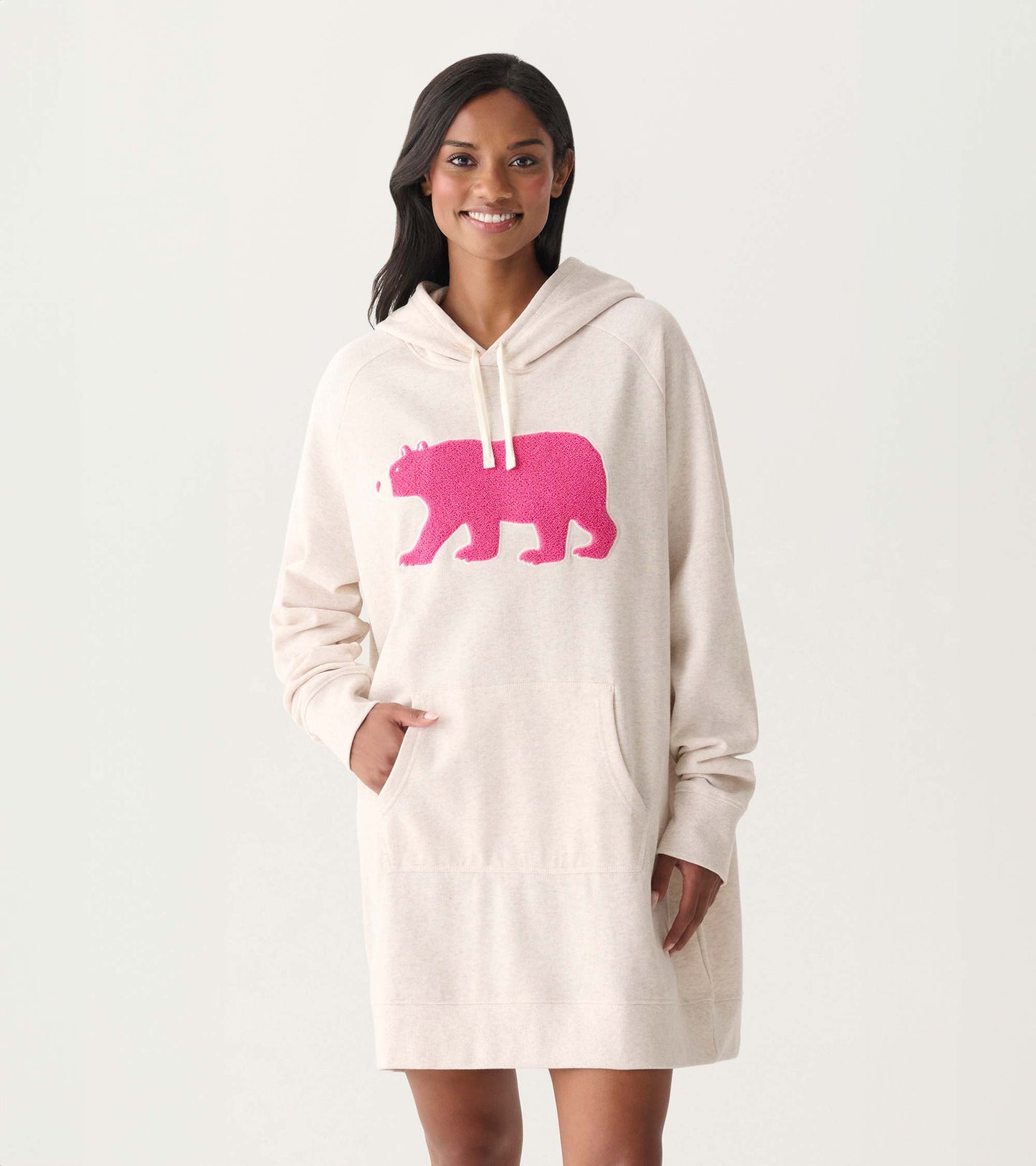Woman wearing an oversized cream hoodie dress with a bright pink bear graphic and front pocket, front view