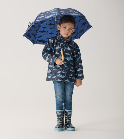 Child wearing dark blue shark print raincoat with matching boots, holding open shark print umbrella