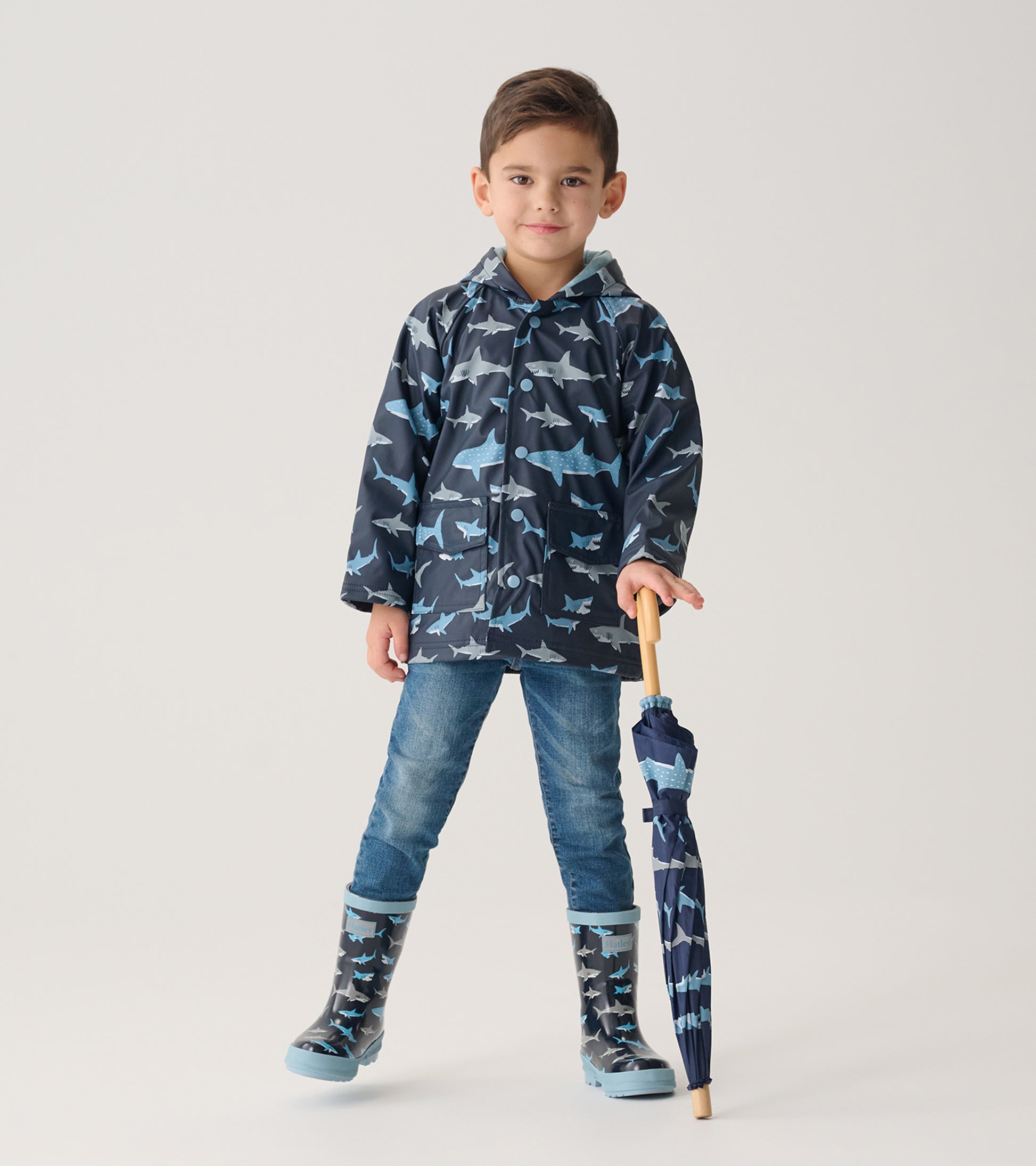 Child wearing dark blue shark print raincoat with matching boots, holding folded umbrella