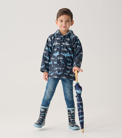 Child wearing dark blue shark print raincoat with matching boots, holding folded umbrella