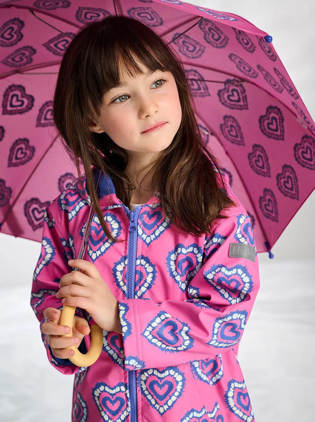 Shop girls' rainwear collection. Showing girl in a pink heart-print raincoat holding a matching umbrella.