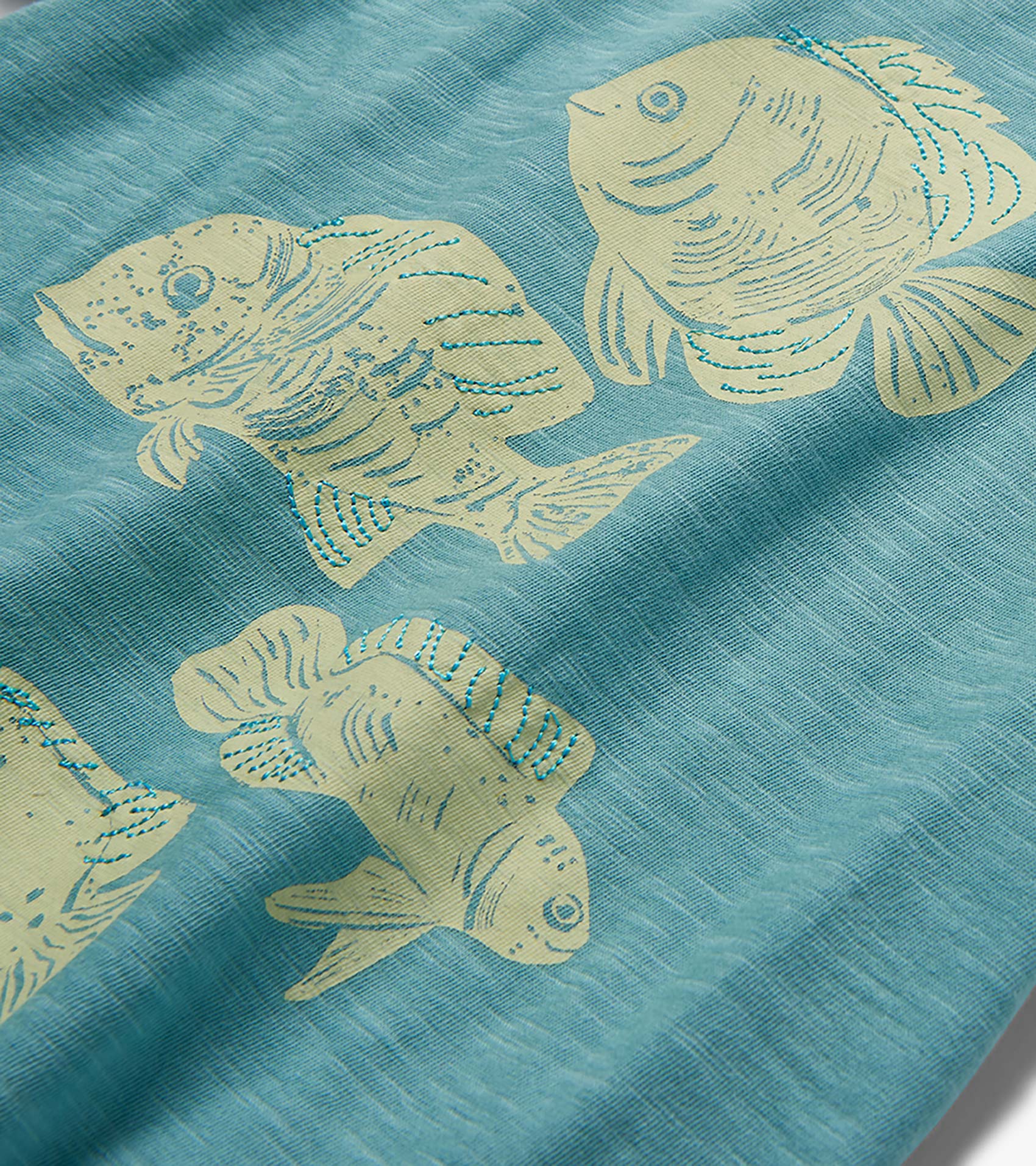 Blue T-shirt fabric with cream fish print and turquoise stitched details, close-up view