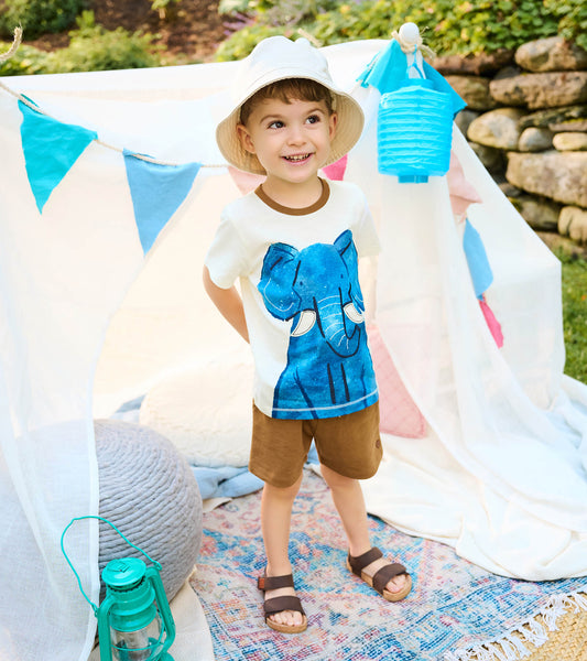 Toddler wearing cream elephant graphic T-shirt with brown shorts, shown standing outdoors